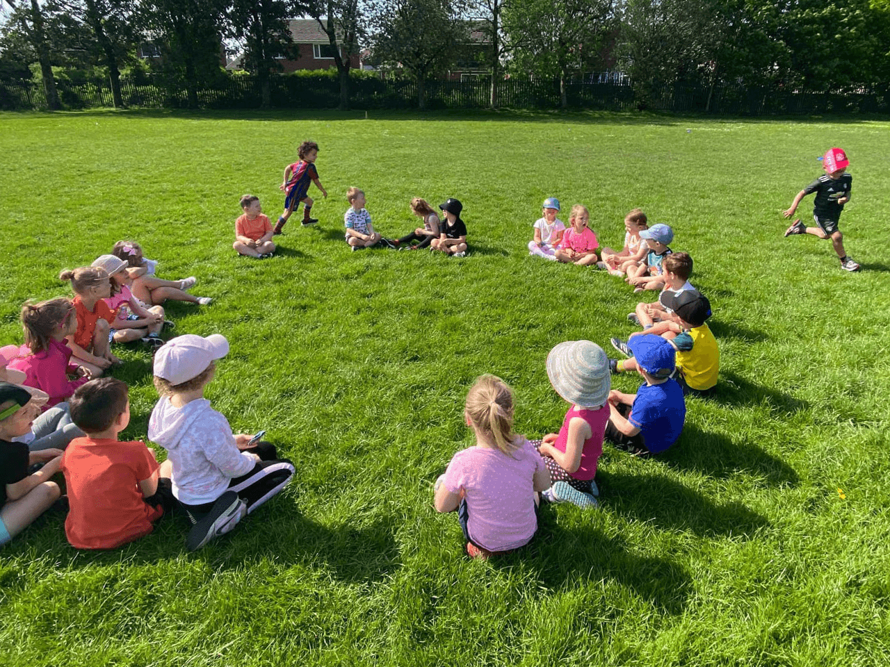 A large group of children sat in a circle on a sports field, playing 'Duck, duck, goose!'