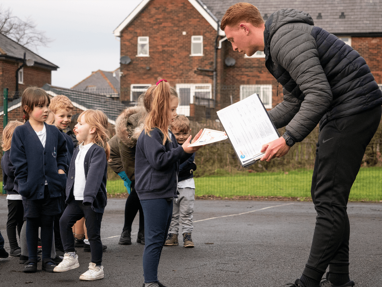 PE coach handing out worksheets to children in the playground.