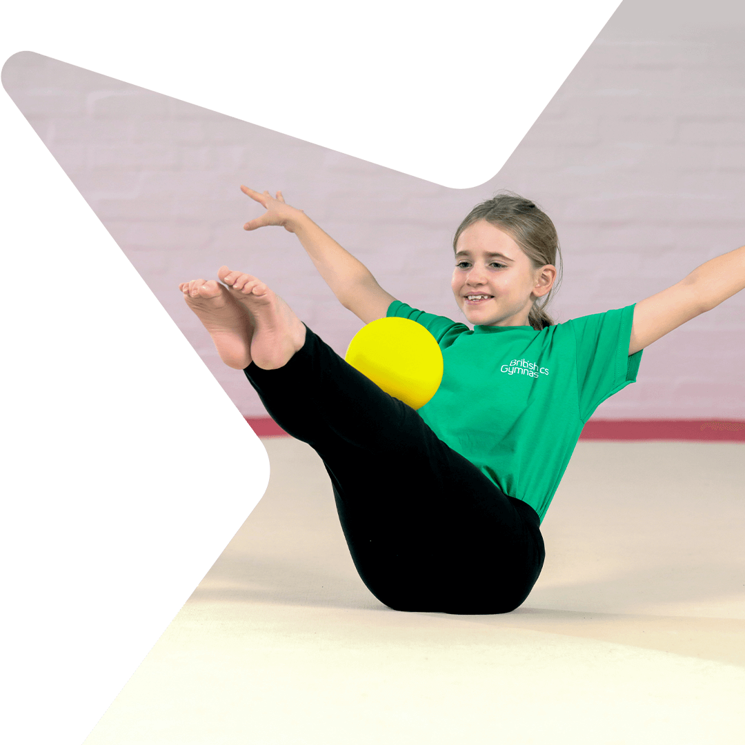 A young female gymnast performing a V-sit balancing a ball on her legs.