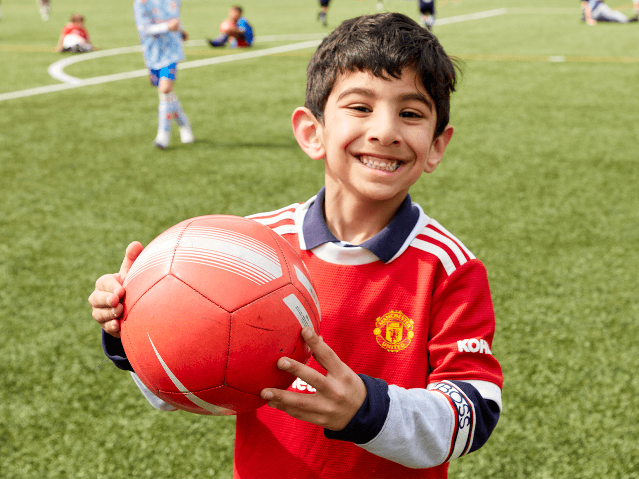 A boy smiling holding a football on a pitch.