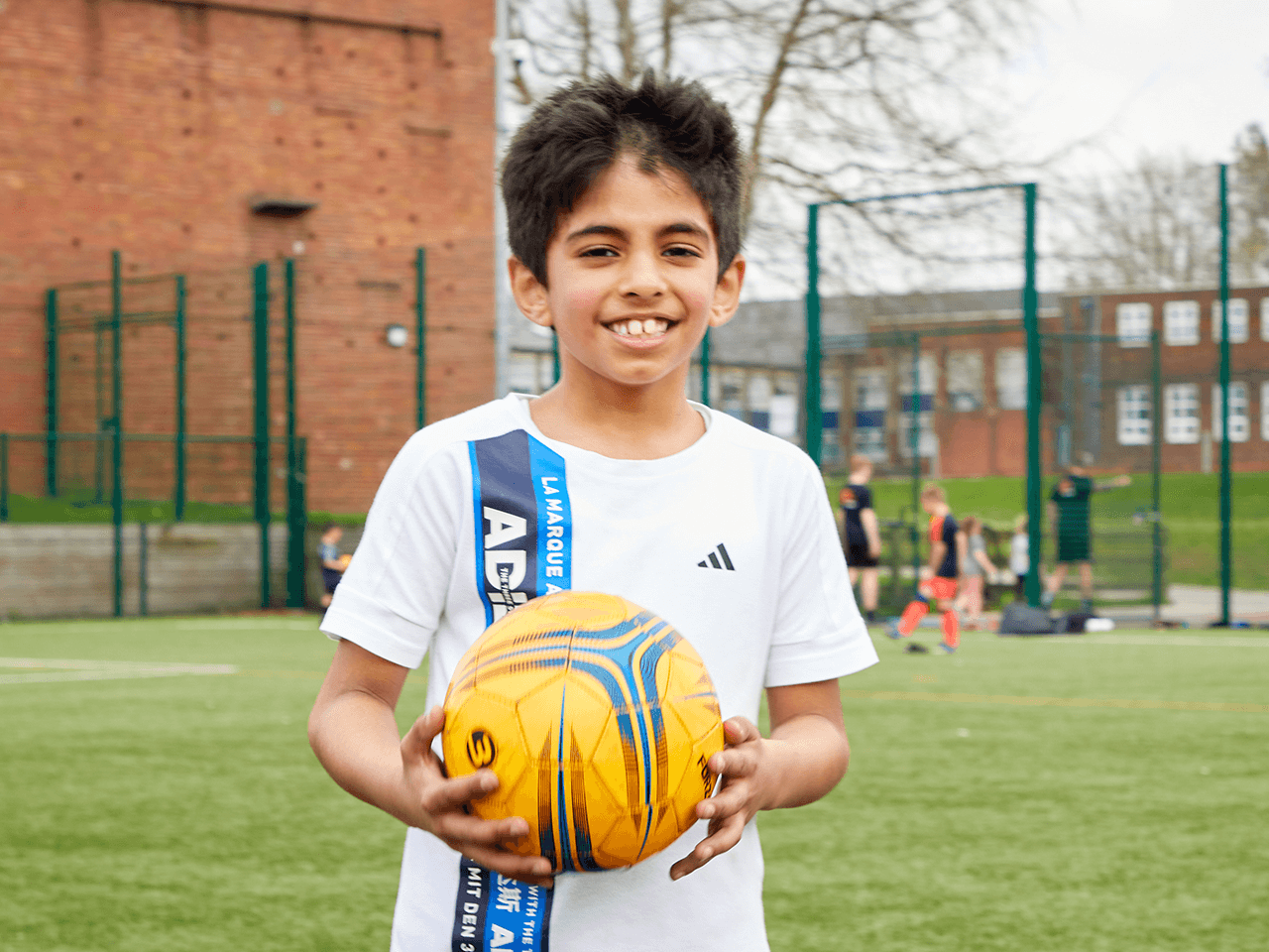 A boy smiling holding a football on a pitch.