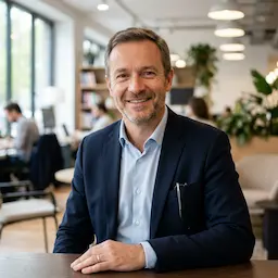 Homme souriant en costume bleu assis à une table dans un bureau moderne avec plantes et lumière naturelle.