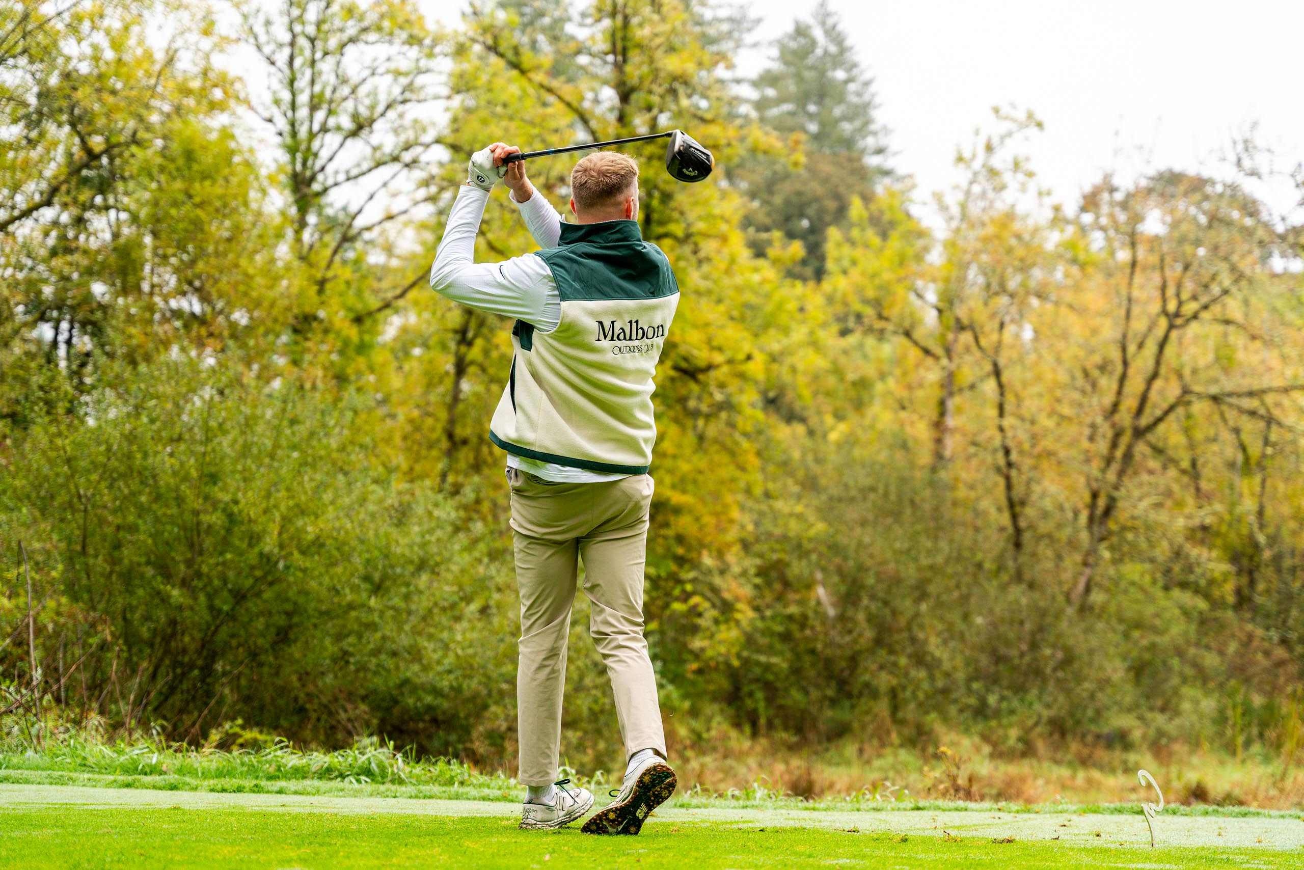Golfer wearing a Malbon Outdoors vest finishing a golf swing on a green course with trees in the background.