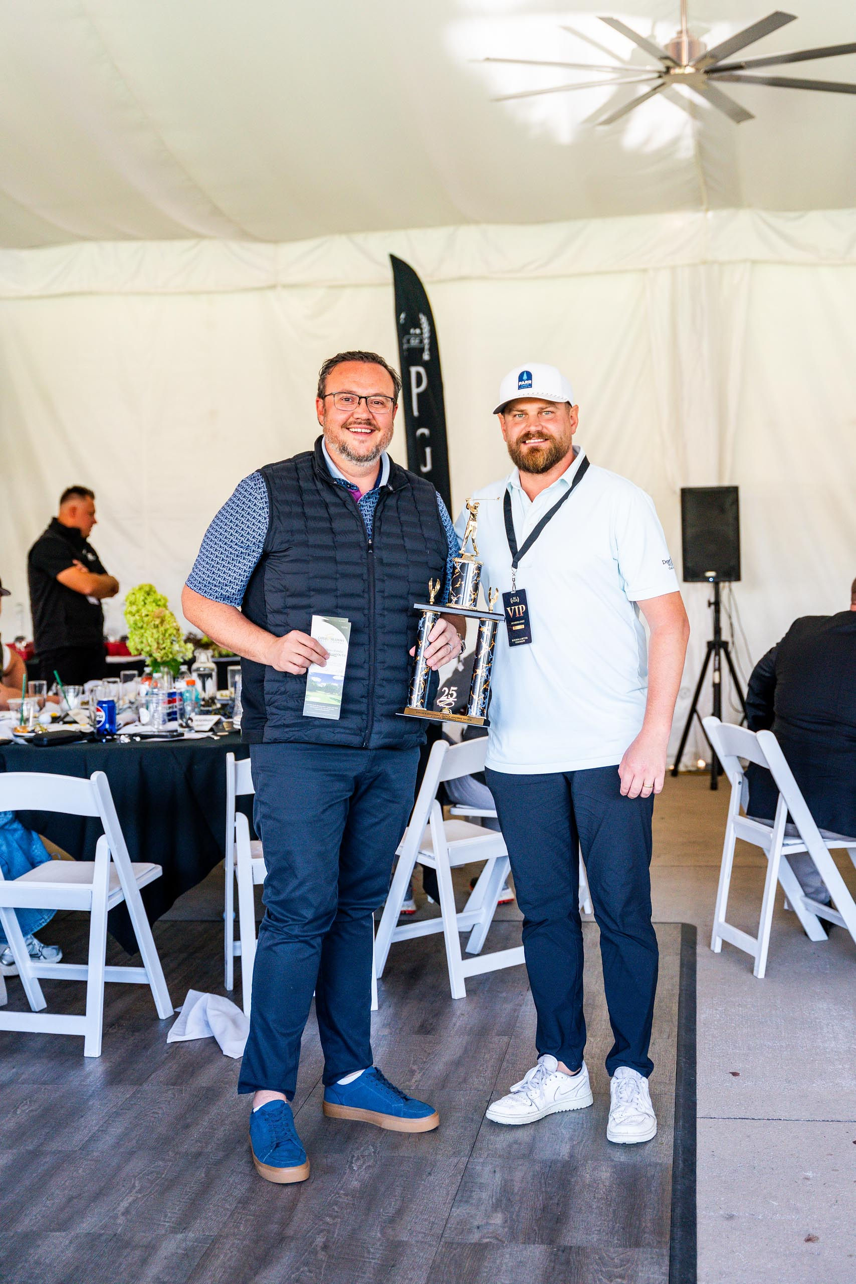 Two men standing indoors, one holding a golf trophy and a brochure, the other wearing a VIP badge and a white cap.