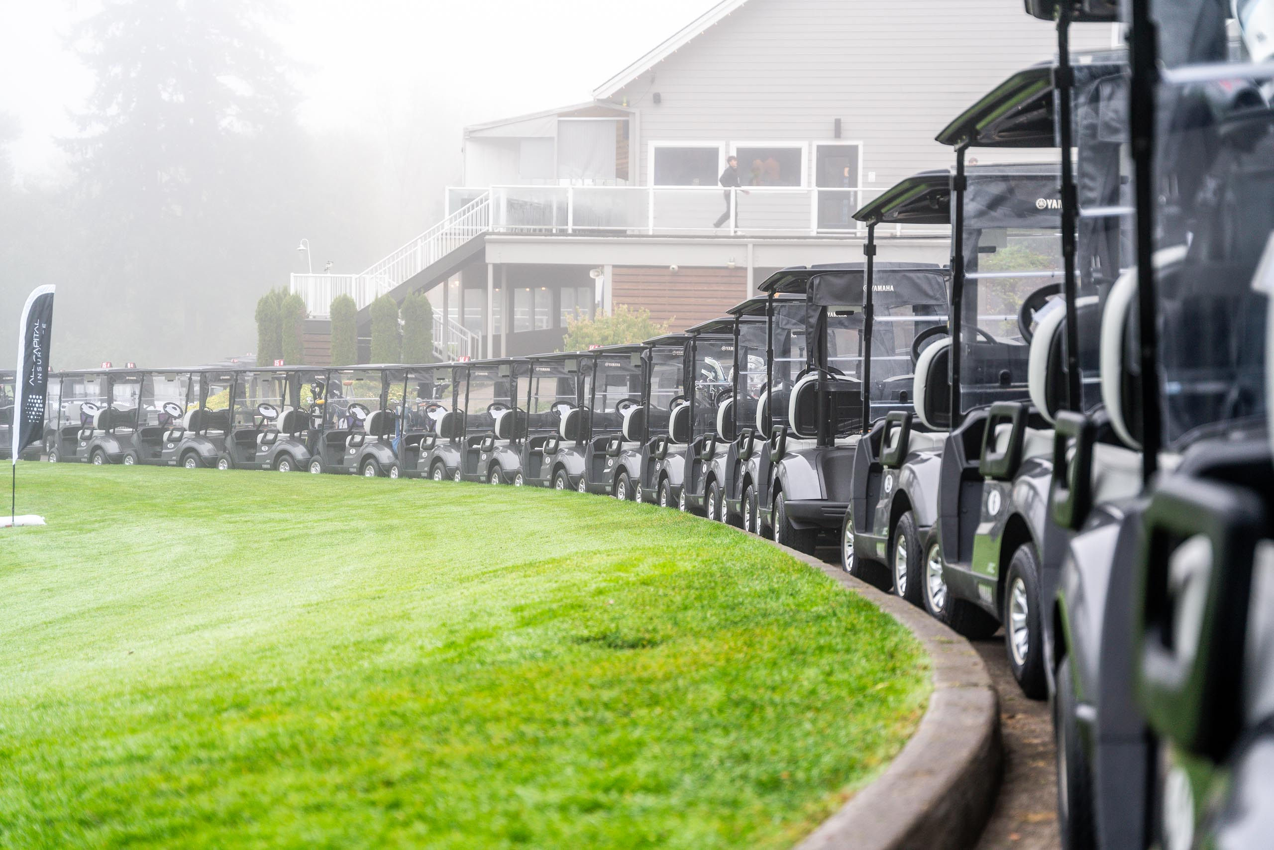 Row of black golf carts lined up on a path beside a green lawn with a building and foggy background.