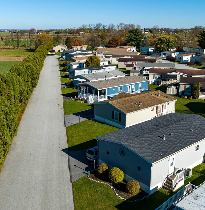 Aerial view of a mobile home park with several rows of colorful manufactured houses along a paved road bordered by tall green trees.