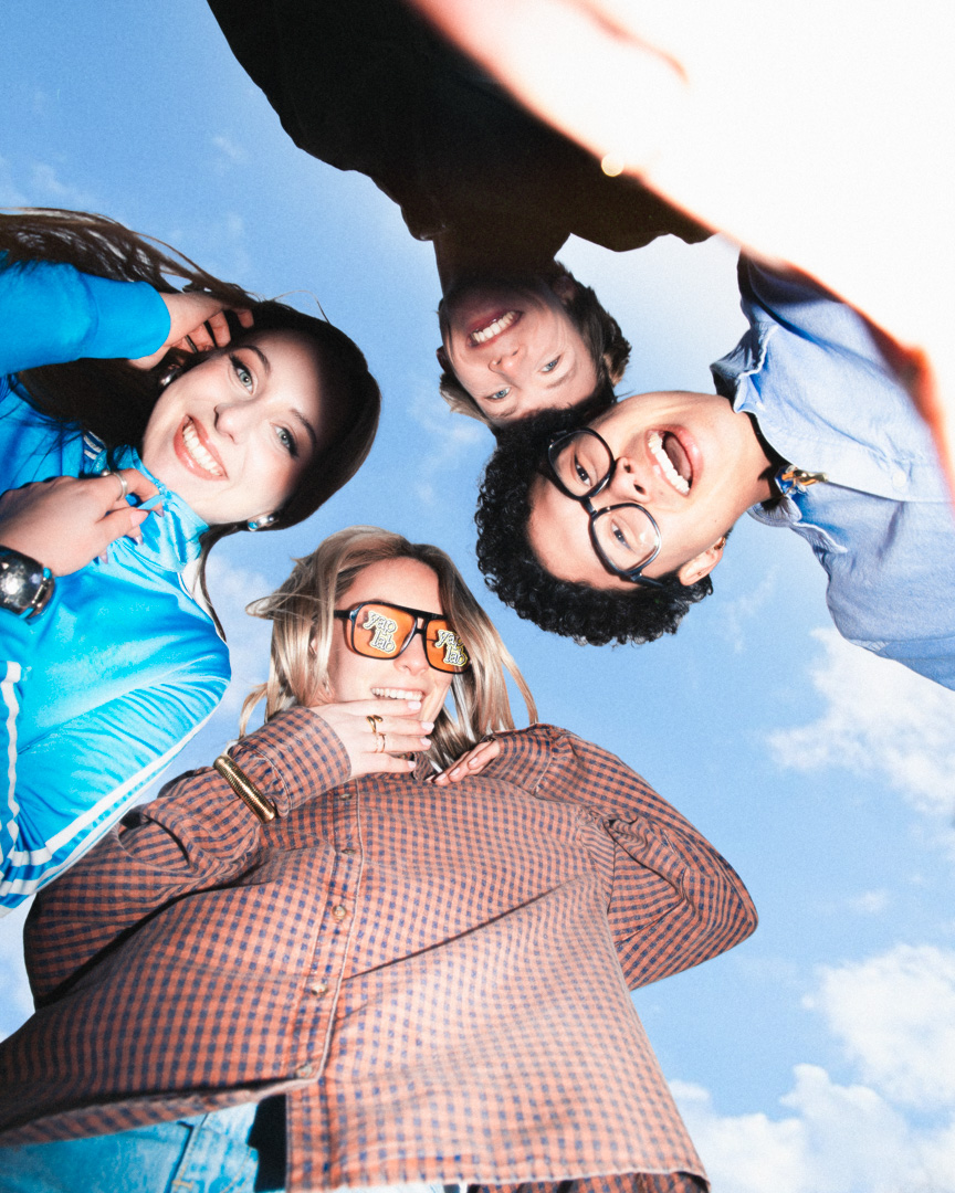 Vier jonge mensen lachen en kijken naar de camera terwijl ze in een cirkel staan met blauwe lucht op de achtergrond.