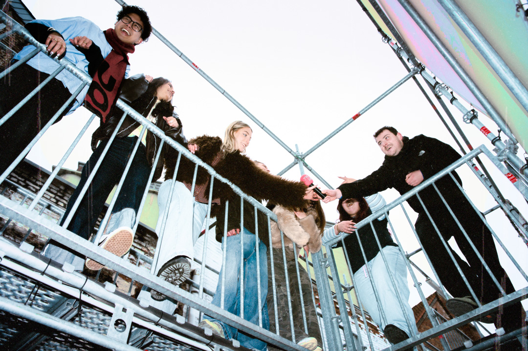 Zes jonge mensen communiceren boven op een metalen steiger, eentje reikt een microfoon aan een ander over de reling.