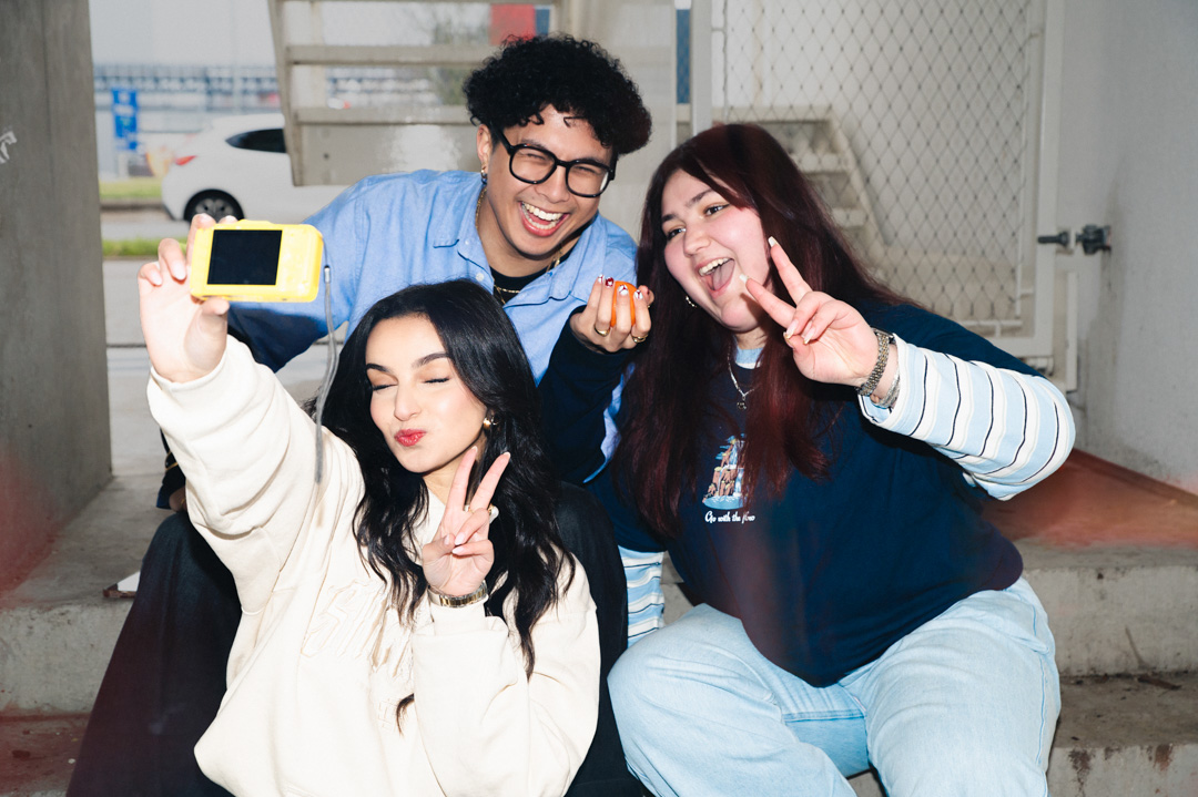 Drie lachende jongeren poseren met een gele camera en maken een peace-teken op een trap.