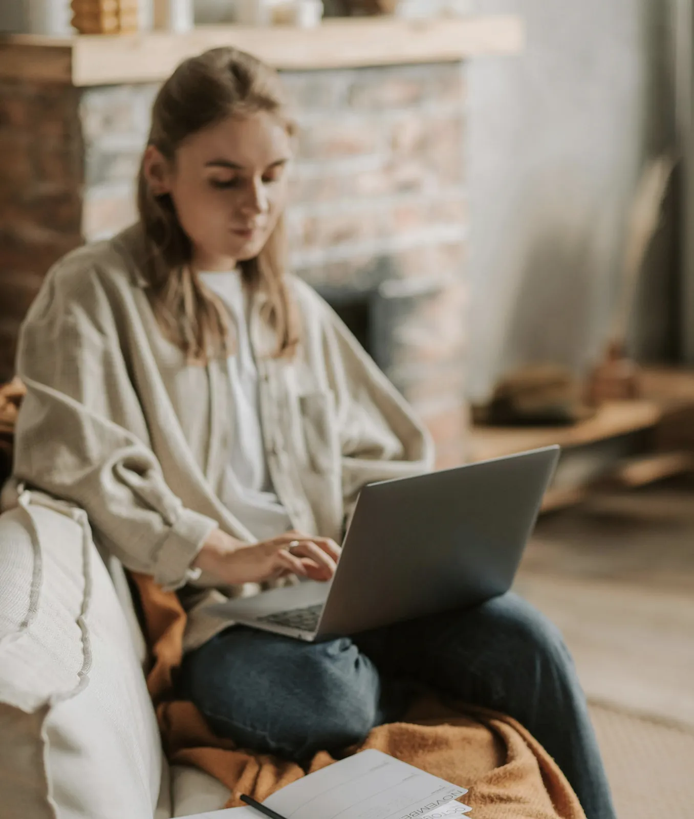 Young woman sitting cross-legged on a couch, working on a laptop with a notebook and pen nearby.