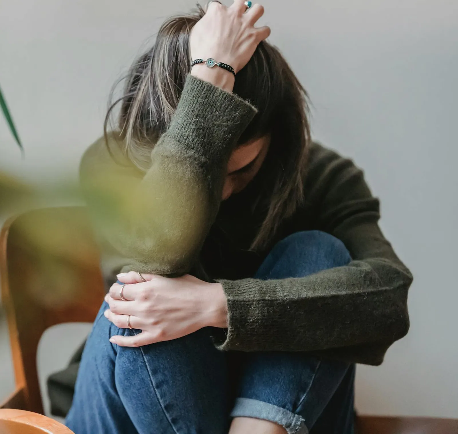 Person sitting with head down, hugging knees and holding their hair with one hand, wearing a green sweater and blue jeans.