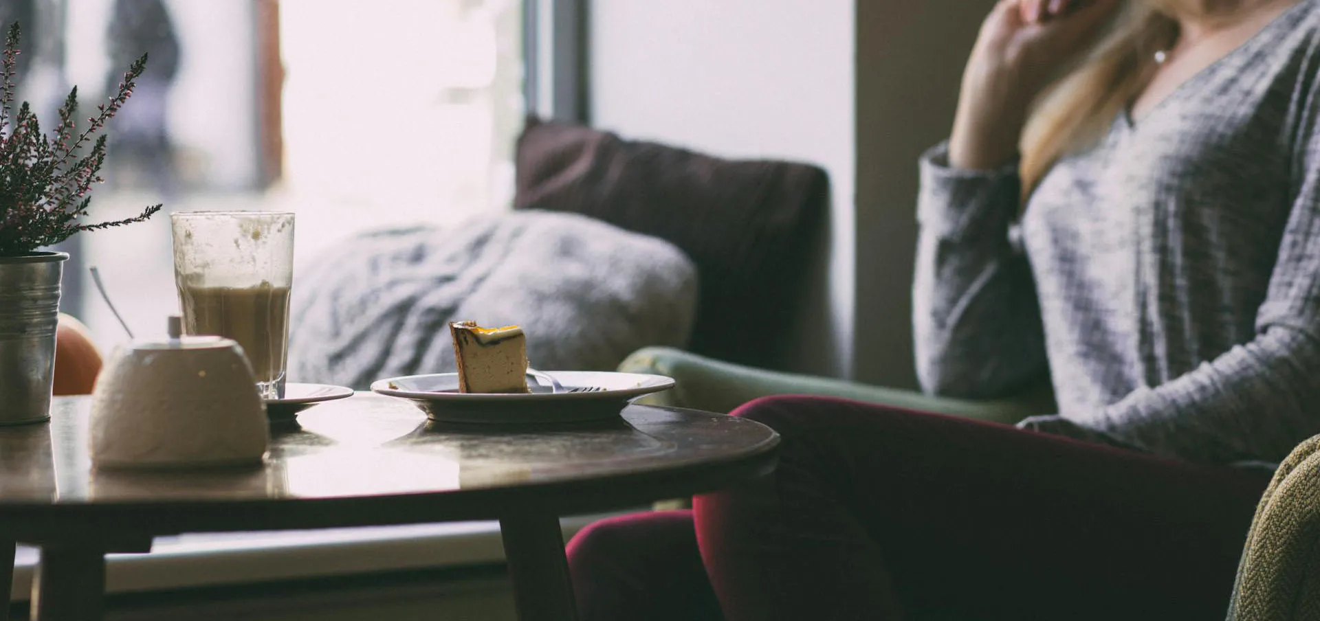Woman sitting indoors near a window with a plate of cake and a glass of coffee on a wooden table.