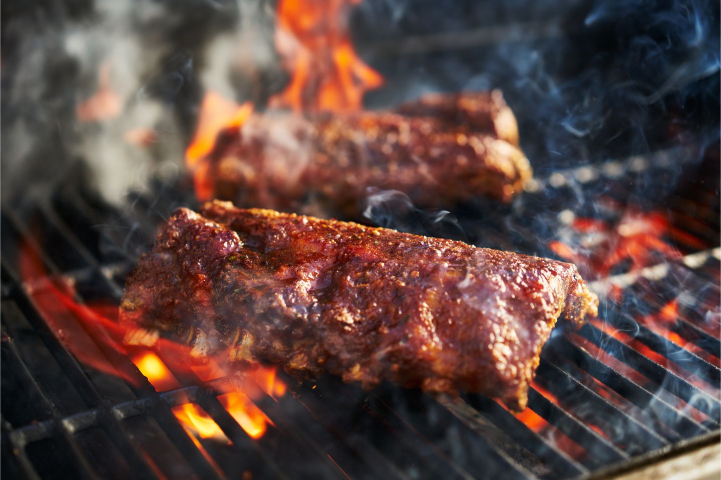 Two racks of seasoned ribs cooking with flames and smoke on a barbecue grill.