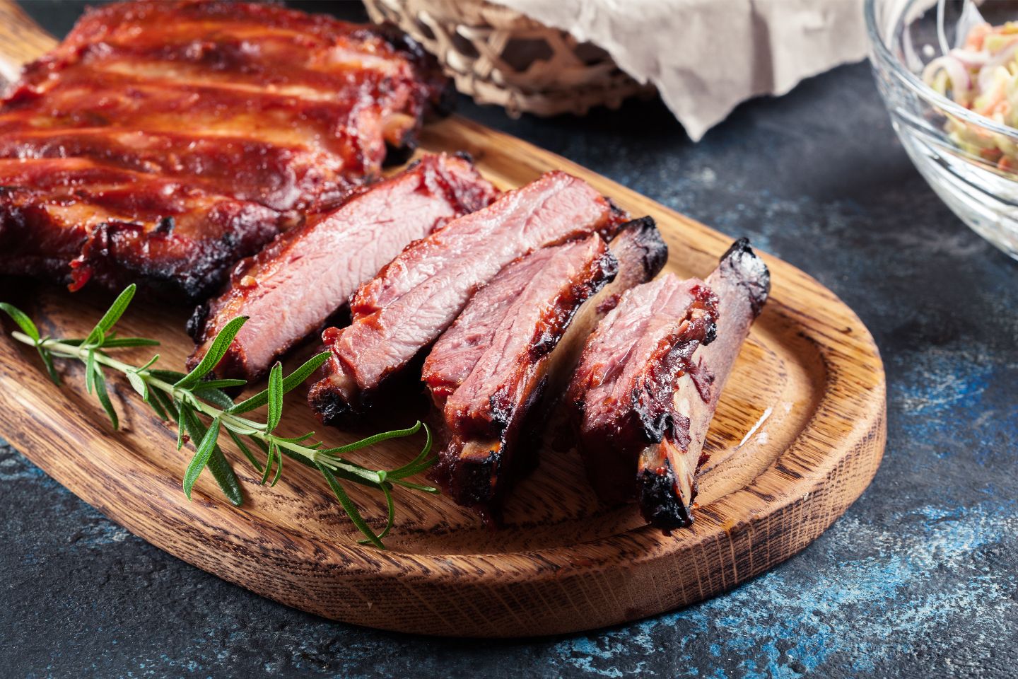 Sliced smoked barbecue ribs with a sprig of rosemary on a wooden serving board.