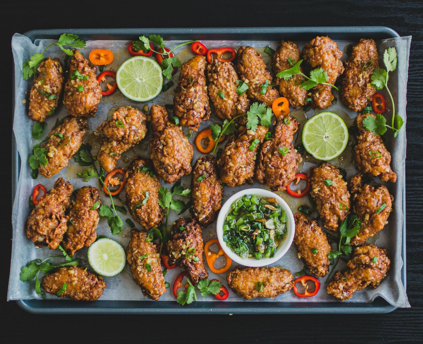 Tray of crispy fried chicken wings garnished with sliced red chili, cilantro, lime halves, and a small bowl of green onion dipping sauce.