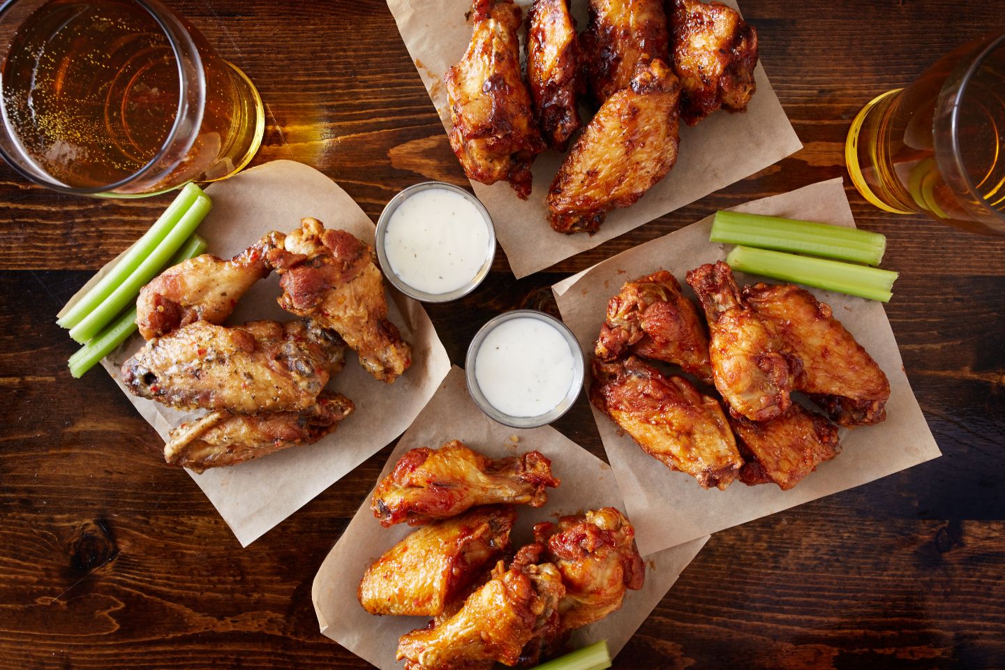 Four servings of chicken wings with celery sticks and two cups of dipping sauce on a wooden table, accompanied by glasses of beer.