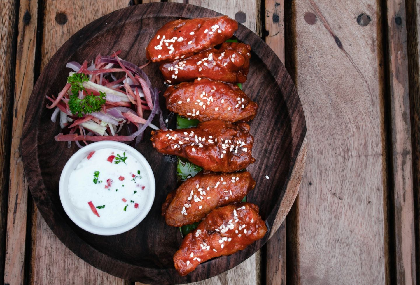Plate of six glazed chicken wings sprinkled with sesame seeds, served with a creamy dipping sauce and a side of sliced vegetables garnished with parsley.
