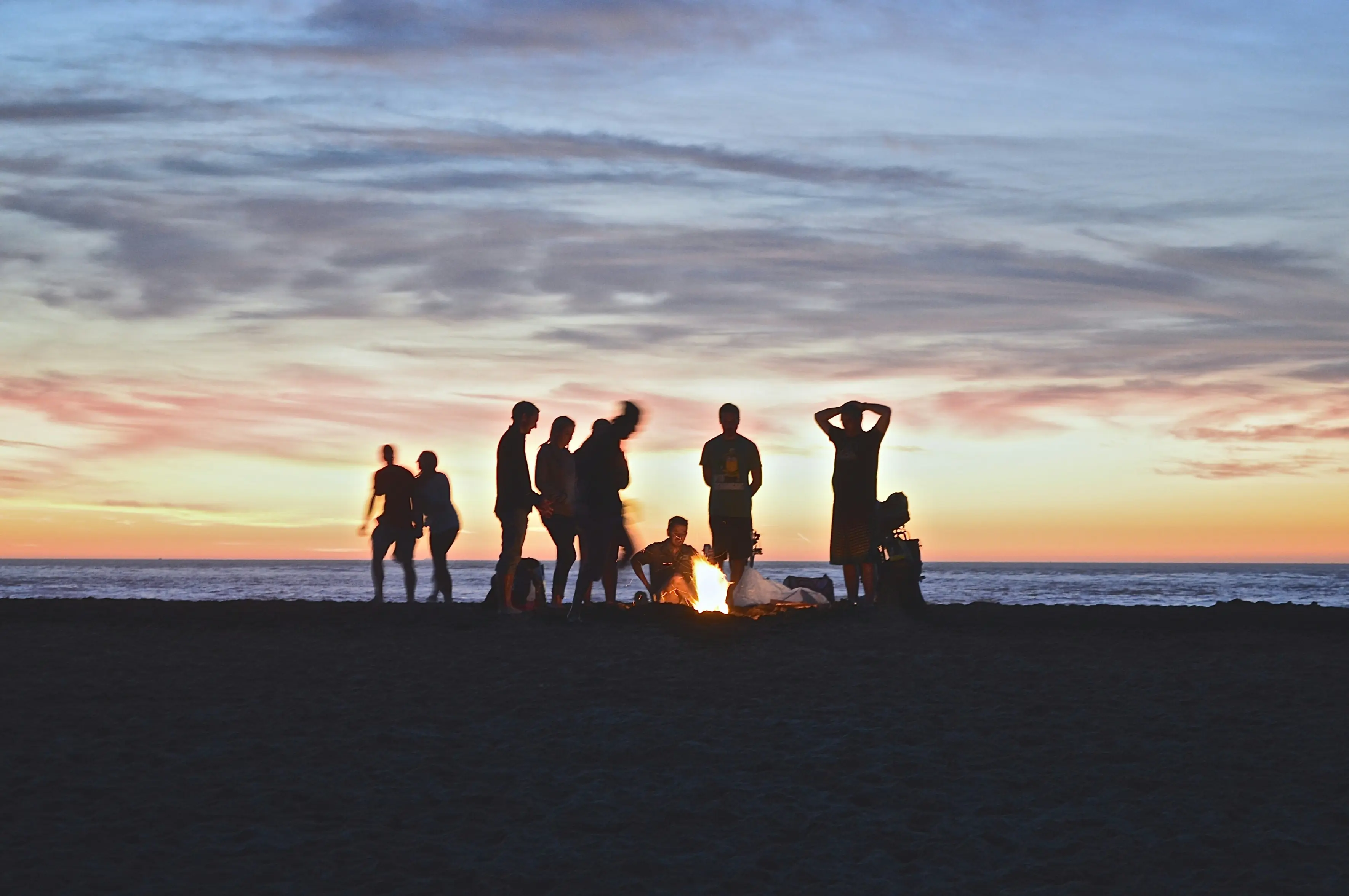 Image of people standing around a beautiful campfire with a sunset behind them. The people are silhouetted.
