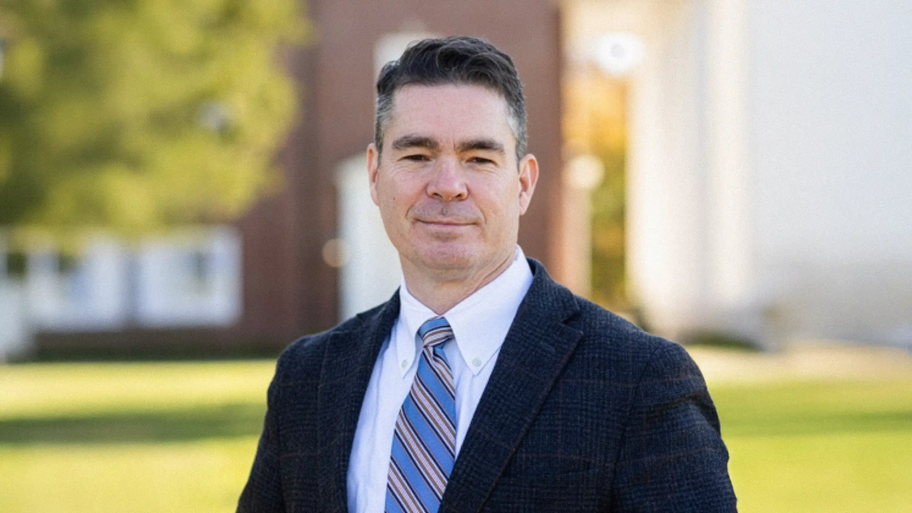 Man with dark hair wearing a blue striped tie and dark blazer standing outdoors with blurred buildings and greenery in the background.