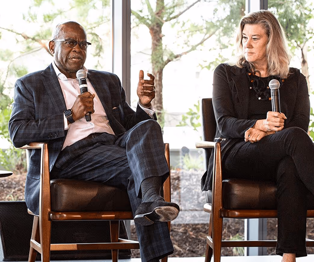 Two people seated on chairs including Dr. Aluko holding microphones, engaged in a discussion in front of large windows with trees outside.