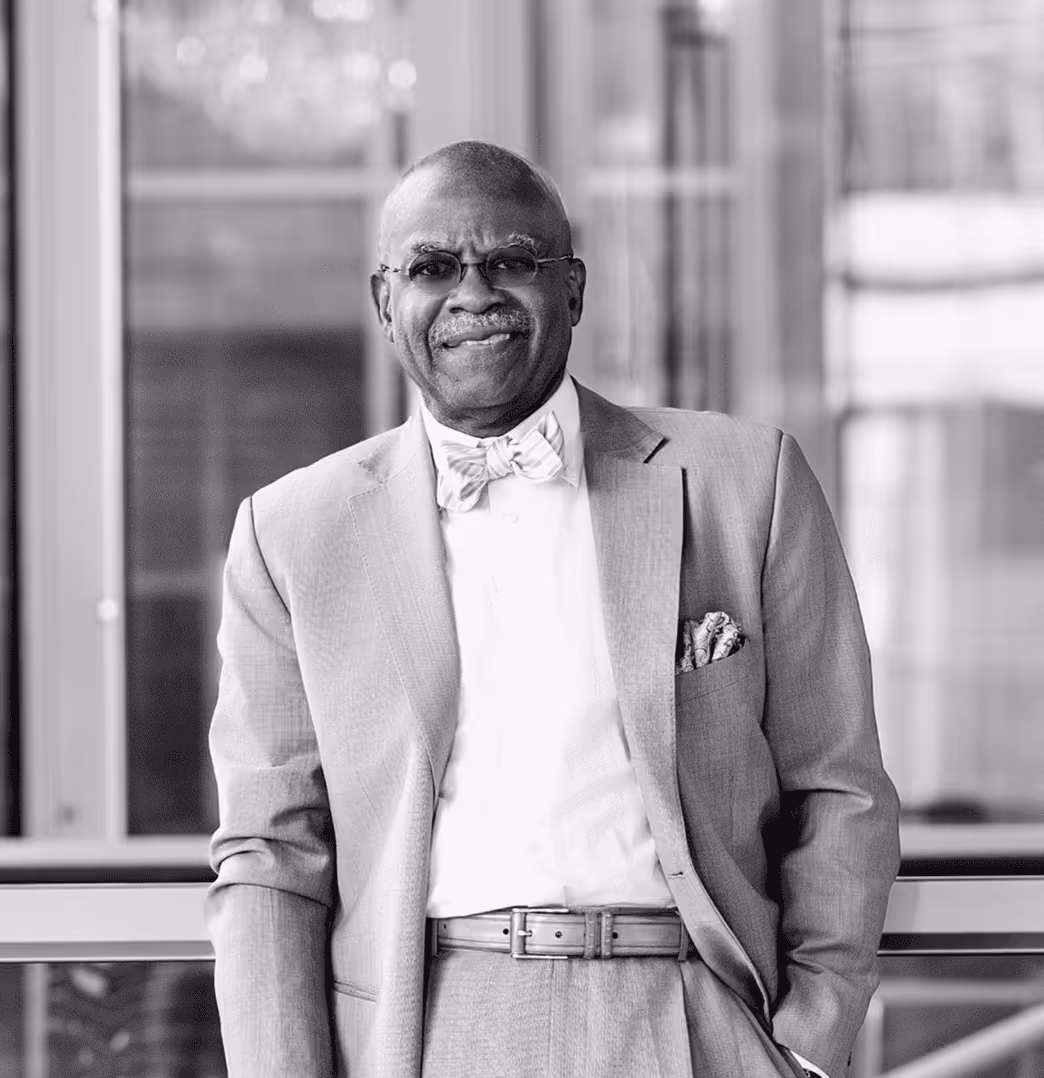 Smiling Dr. Aluko wearing glasses, a bow tie, and a suit with a pocket square, standing indoors.
