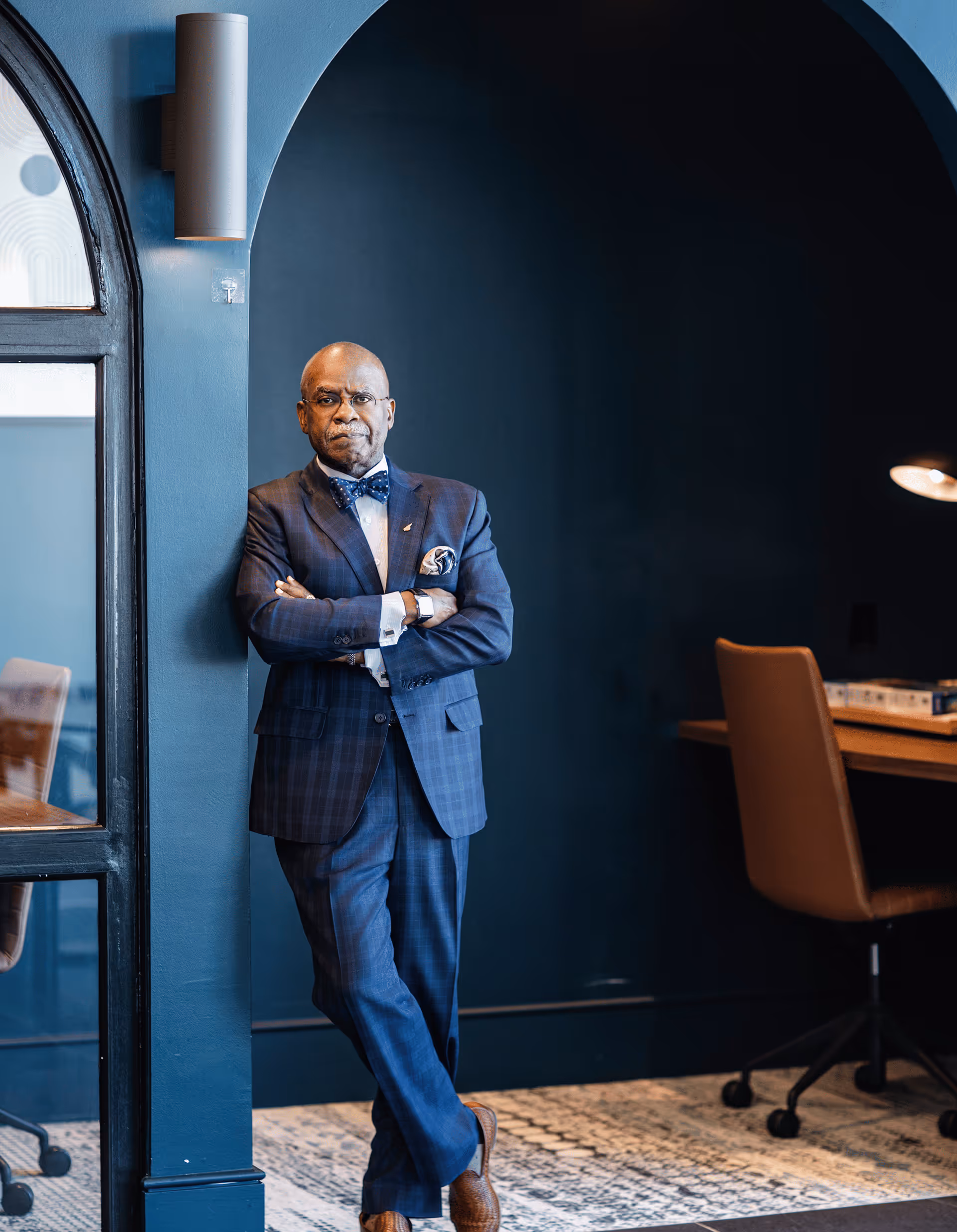 Dr. Aluko in a blue checkered suit with bow tie leaning against a wall in a modern office space.