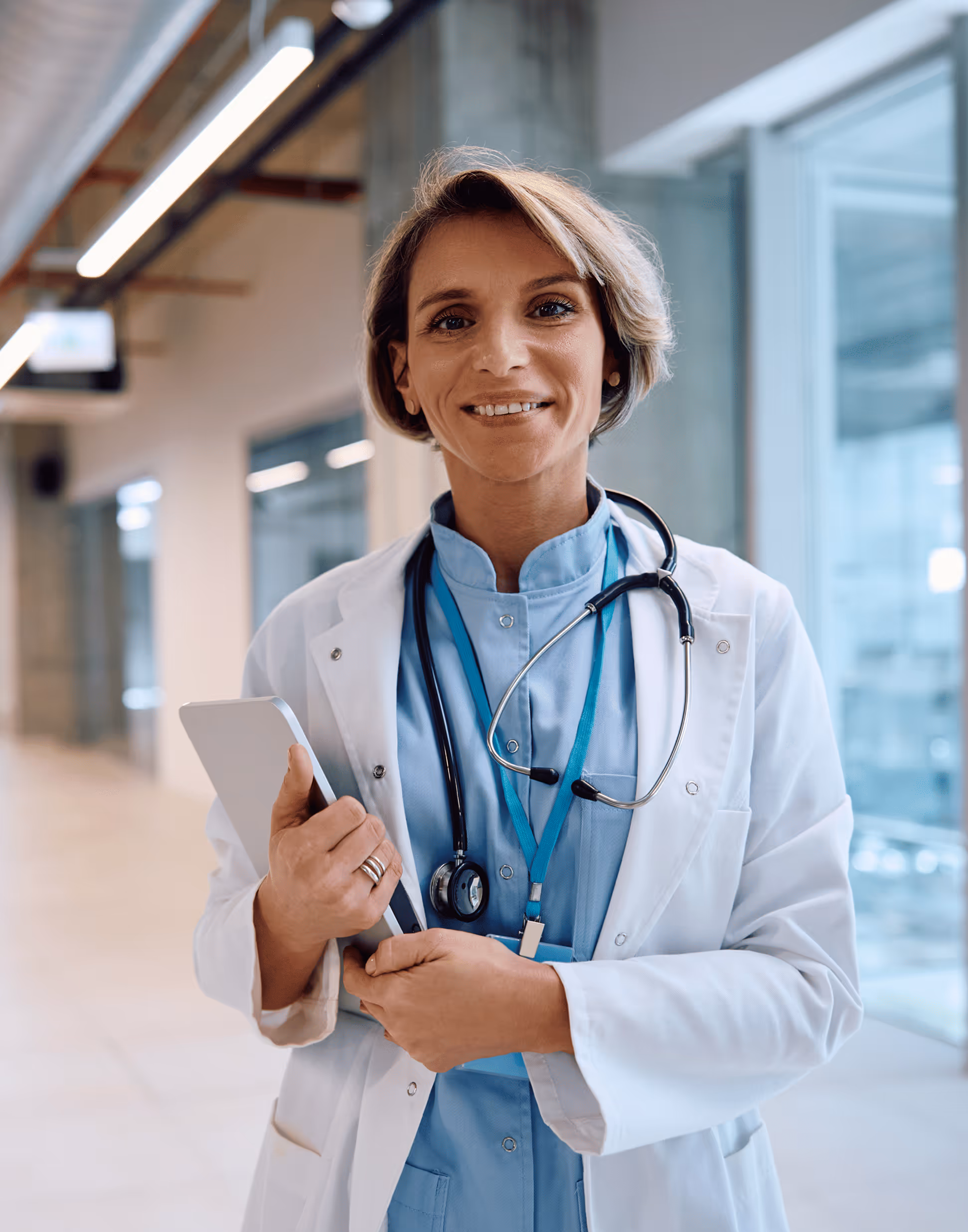 Female doctor in white coat and blue scrubs holding a tablet and smiling in a hospital corridor.
