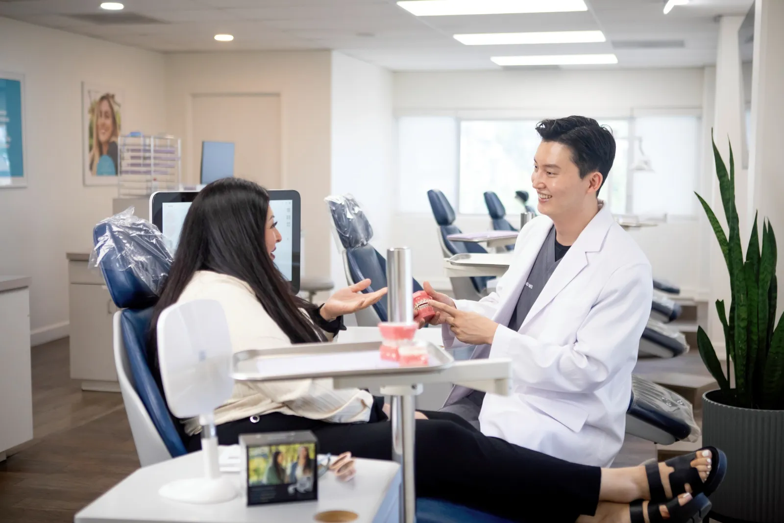 Dentist in white coat showing dental model to a seated female patient in a bright dental office.