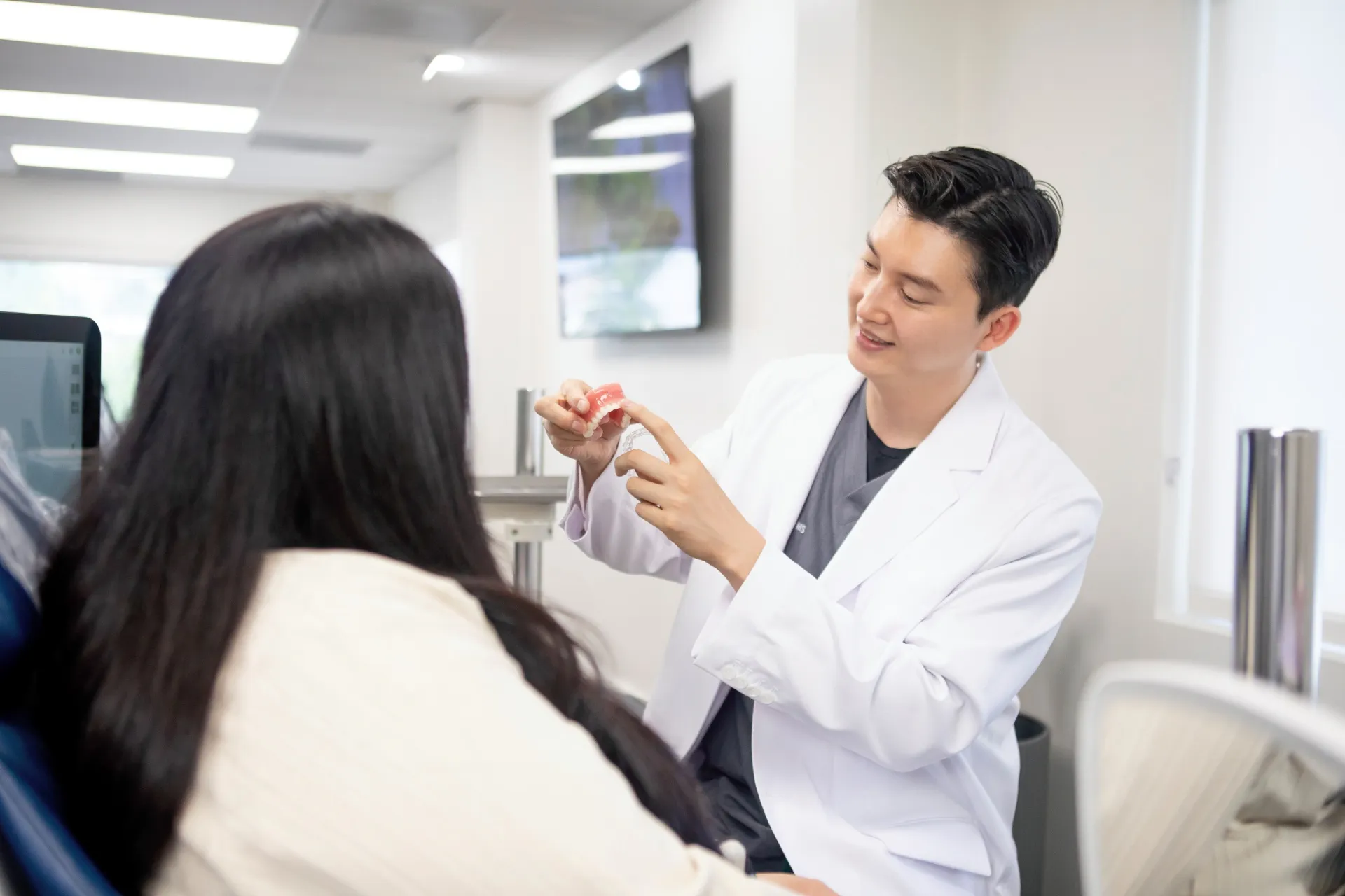Dentist in white coat showing a dental model to a patient with long dark hair in a dental office.