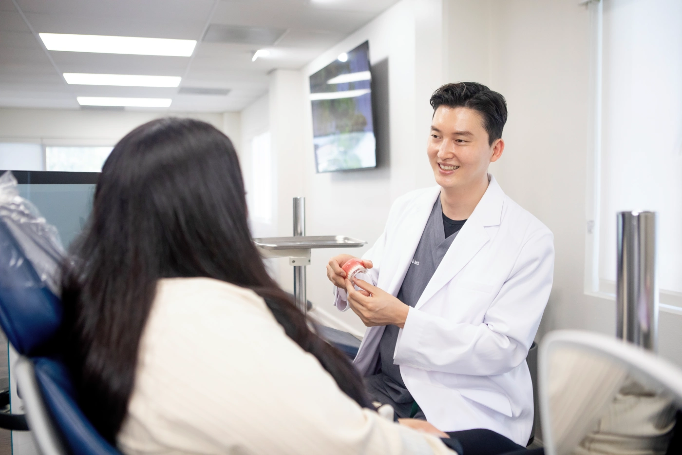 Dentist in a white coat smiling and showing a dental model to a seated patient with long hair in a dental office.