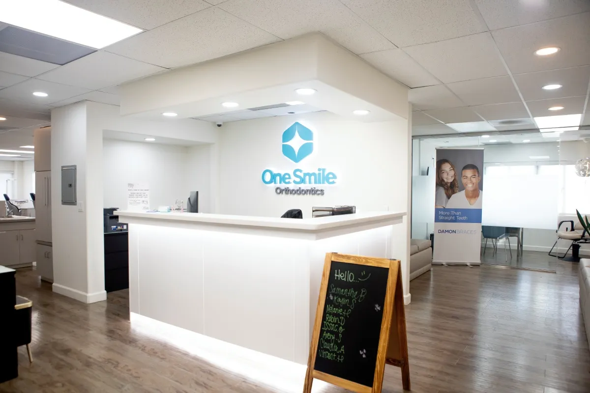 Reception desk inside a modern orthodontic office with illuminated One Smile Orthodontics logo on the wall and a wooden stand-up chalkboard with staff names.