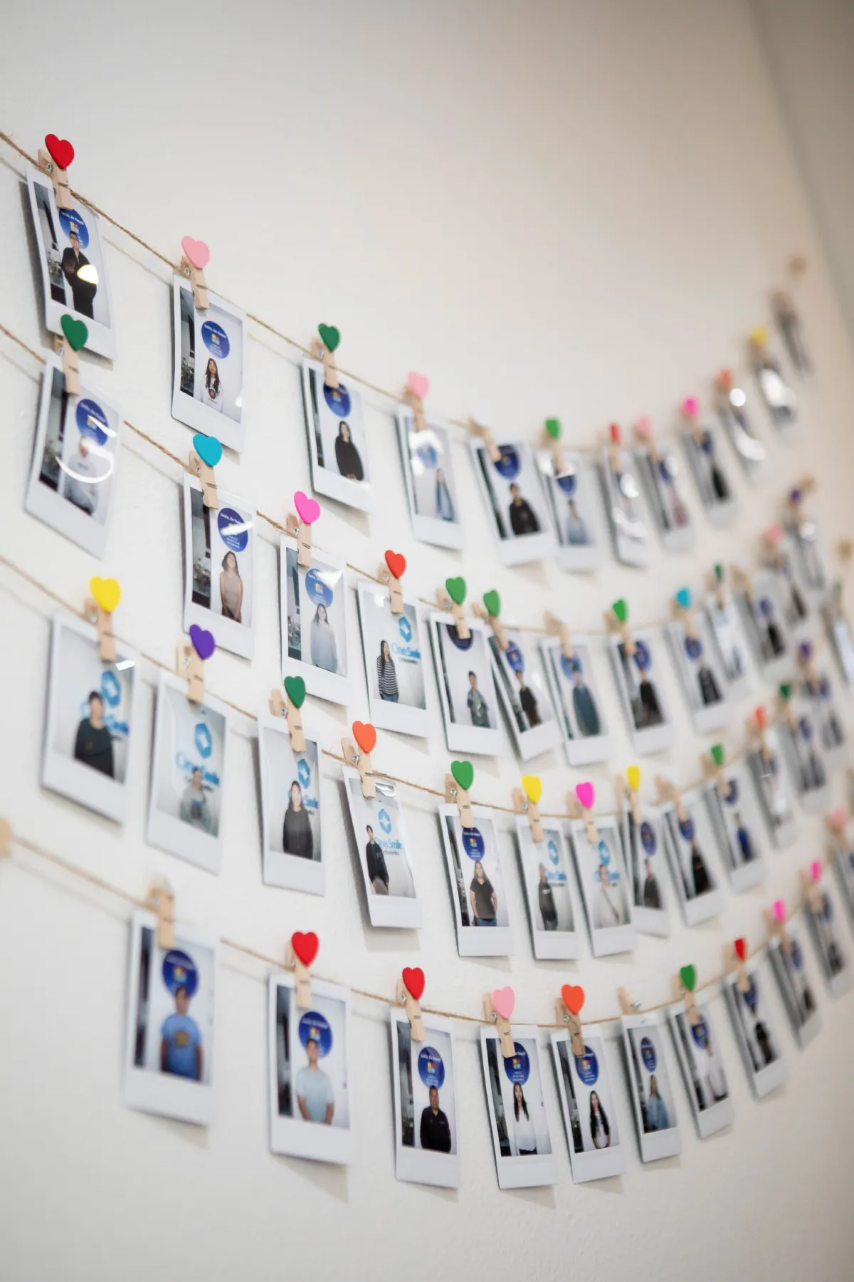 Multiple rows of Polaroid photos clipped with colorful heart-shaped clips on strings hanging on a wall.