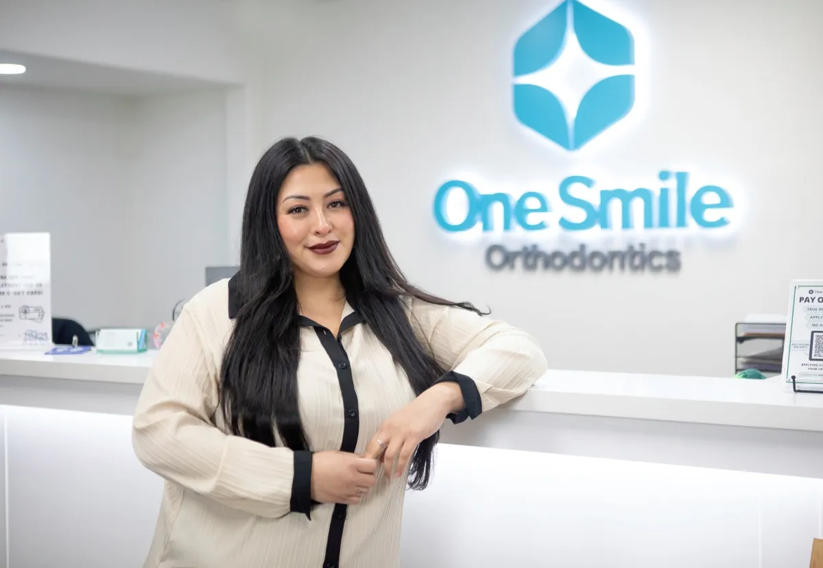 Woman with long black hair and dark lipstick leaning on a white counter in an orthodontics office with One Smile Orthodontics logo on the wall.