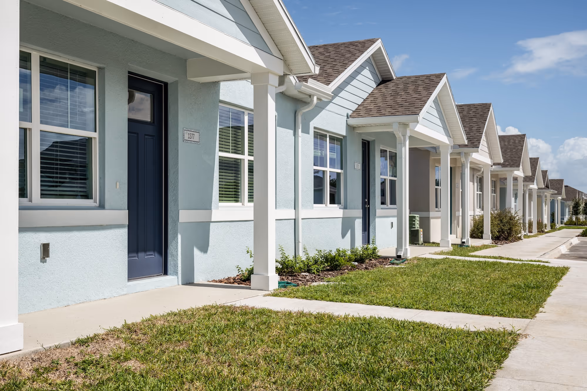 Row of modern single-story townhouses with light blue walls, dark blue doors, white trim, and small front lawns under a clear sky.
