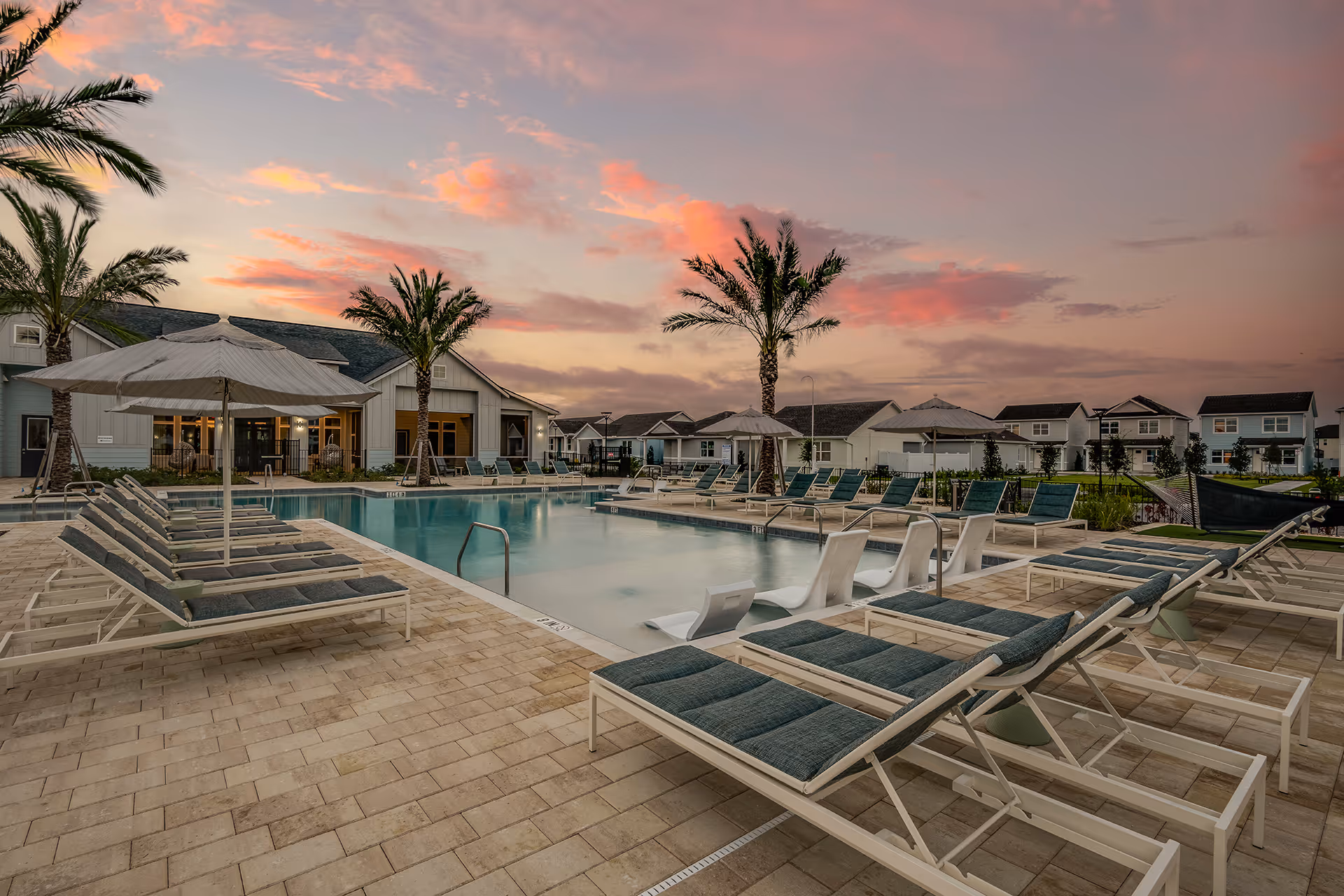 Outdoor pool with lounge chairs and umbrellas at sunset in a residential neighborhood with palm trees.