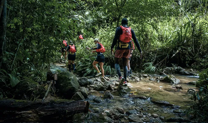 Runners in red packs crossing a rocky jungle stream in single file