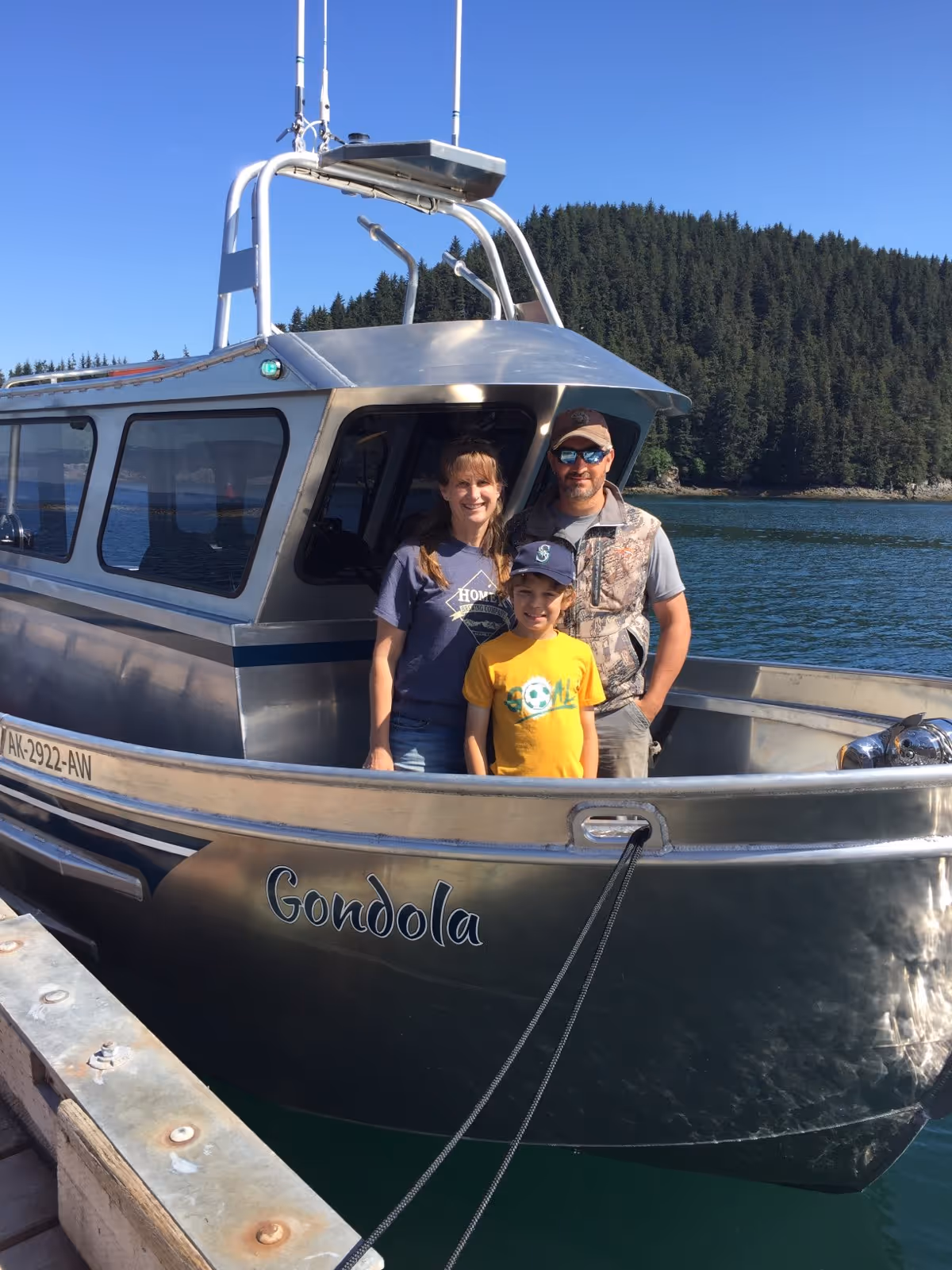 A family of three standing on the deck of a silver boat named Gondola, docked by a wooden pier with forested hills and blue sky in the background.