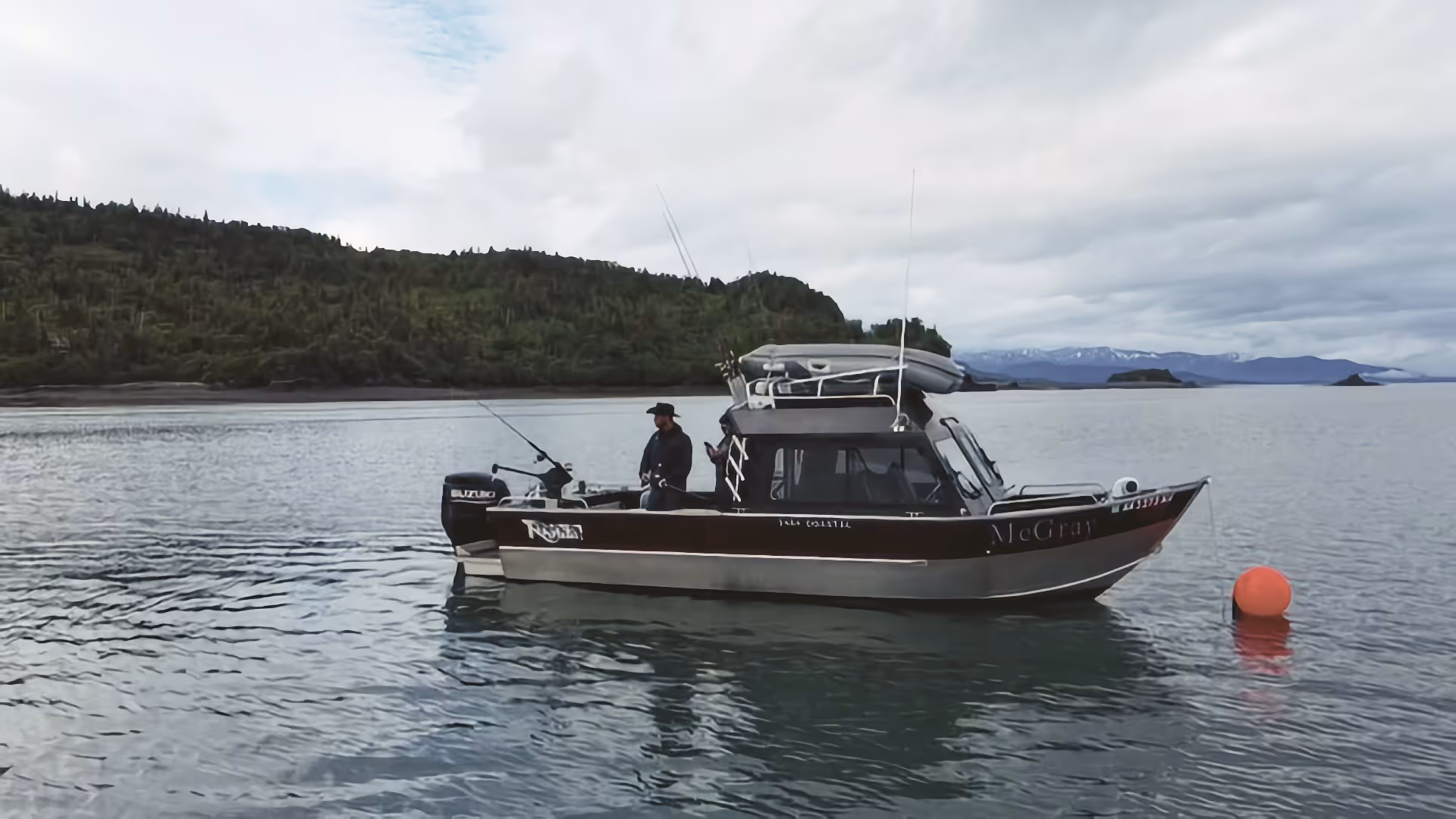 Fishing boat named McGray floating on calm water near a green forested shore under a cloudy sky.