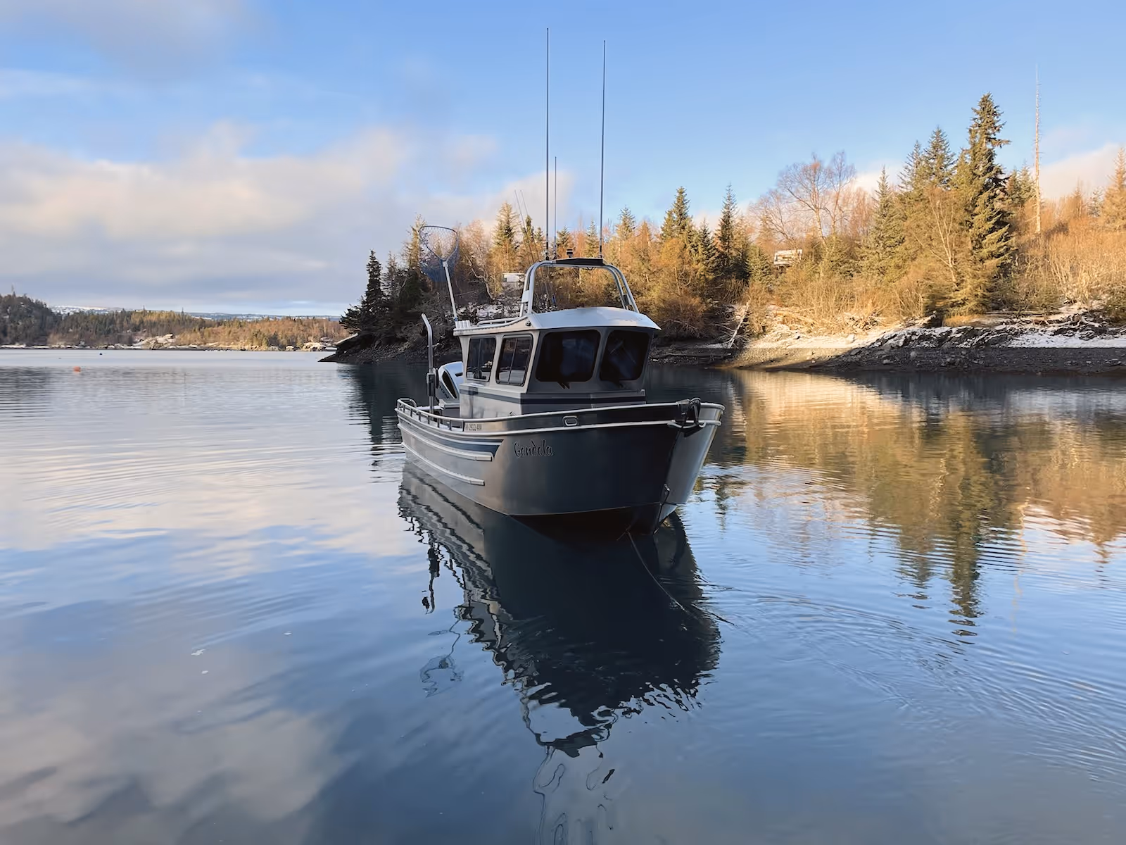 Small fishing boat named Gondola floating on calm water near a shoreline with evergreen trees and a partly cloudy sky.