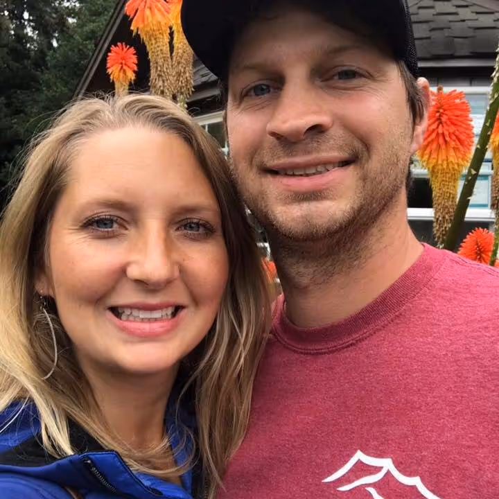 Smiling man and woman posing closely outdoors in front of orange and yellow flowers and a house window.
