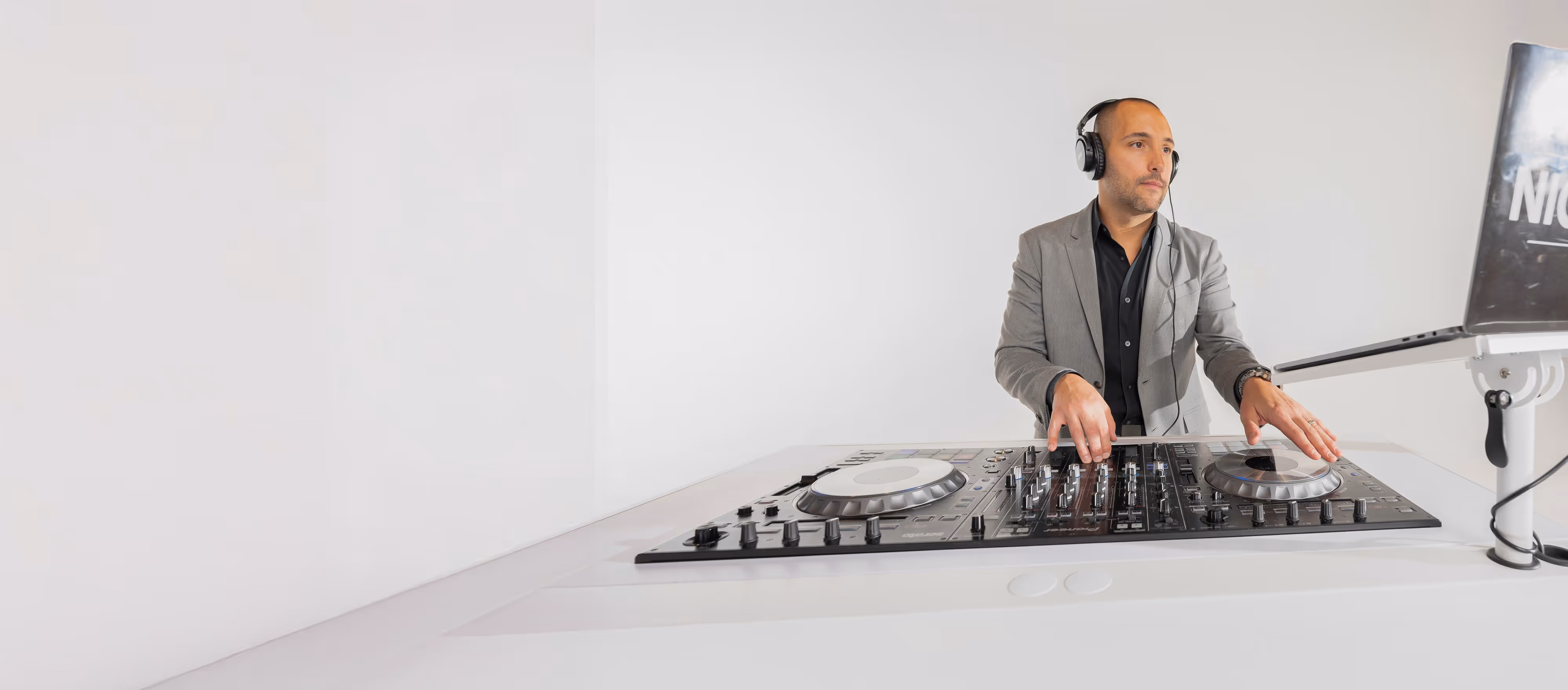 A man wearing headphones and a gray blazer operating a DJ mixer and laptop in a bright, minimal setting.