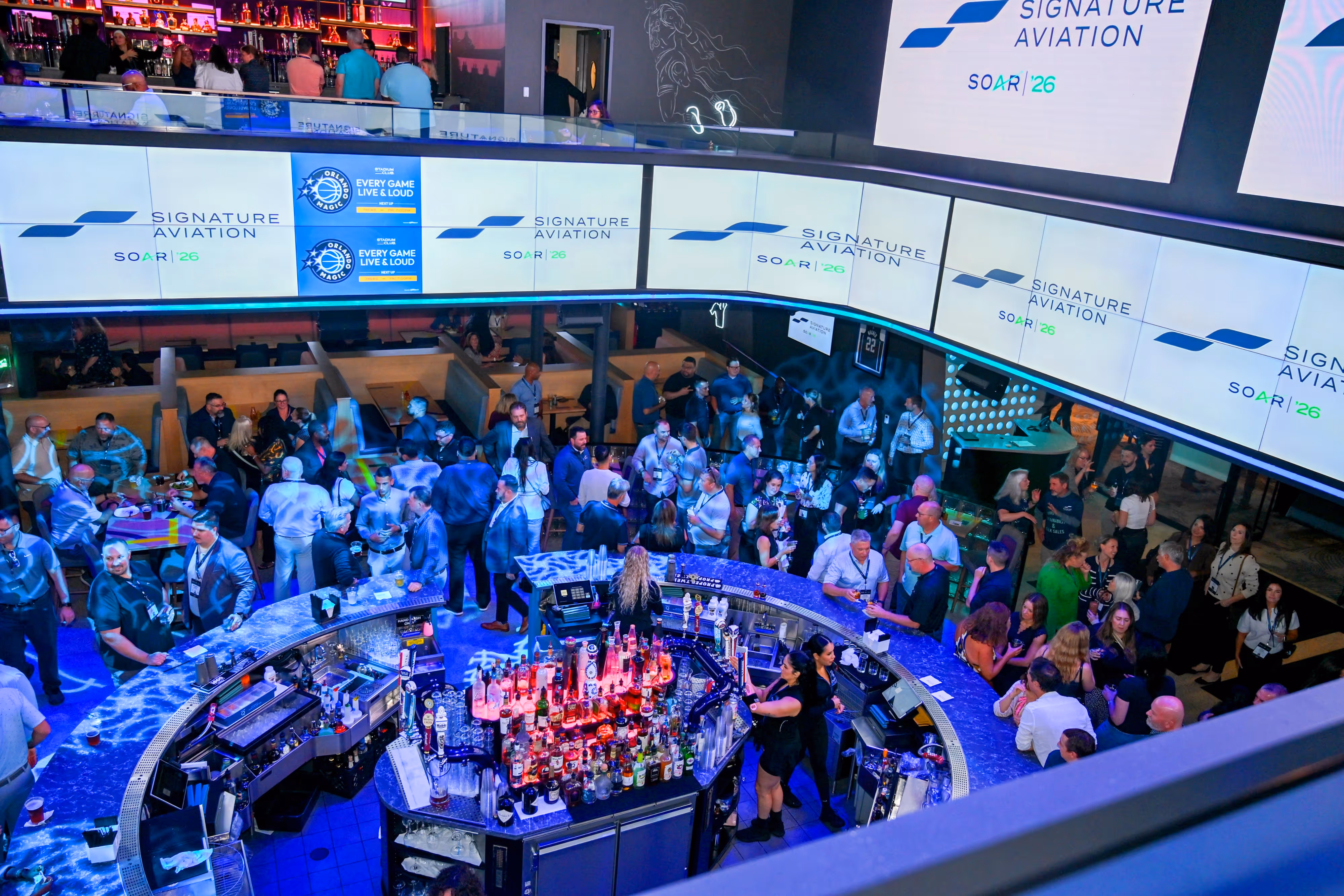 Crowded bar with people socializing around a circular counter stocked with various bottles under large digital screens displaying Signature Aviation logos.