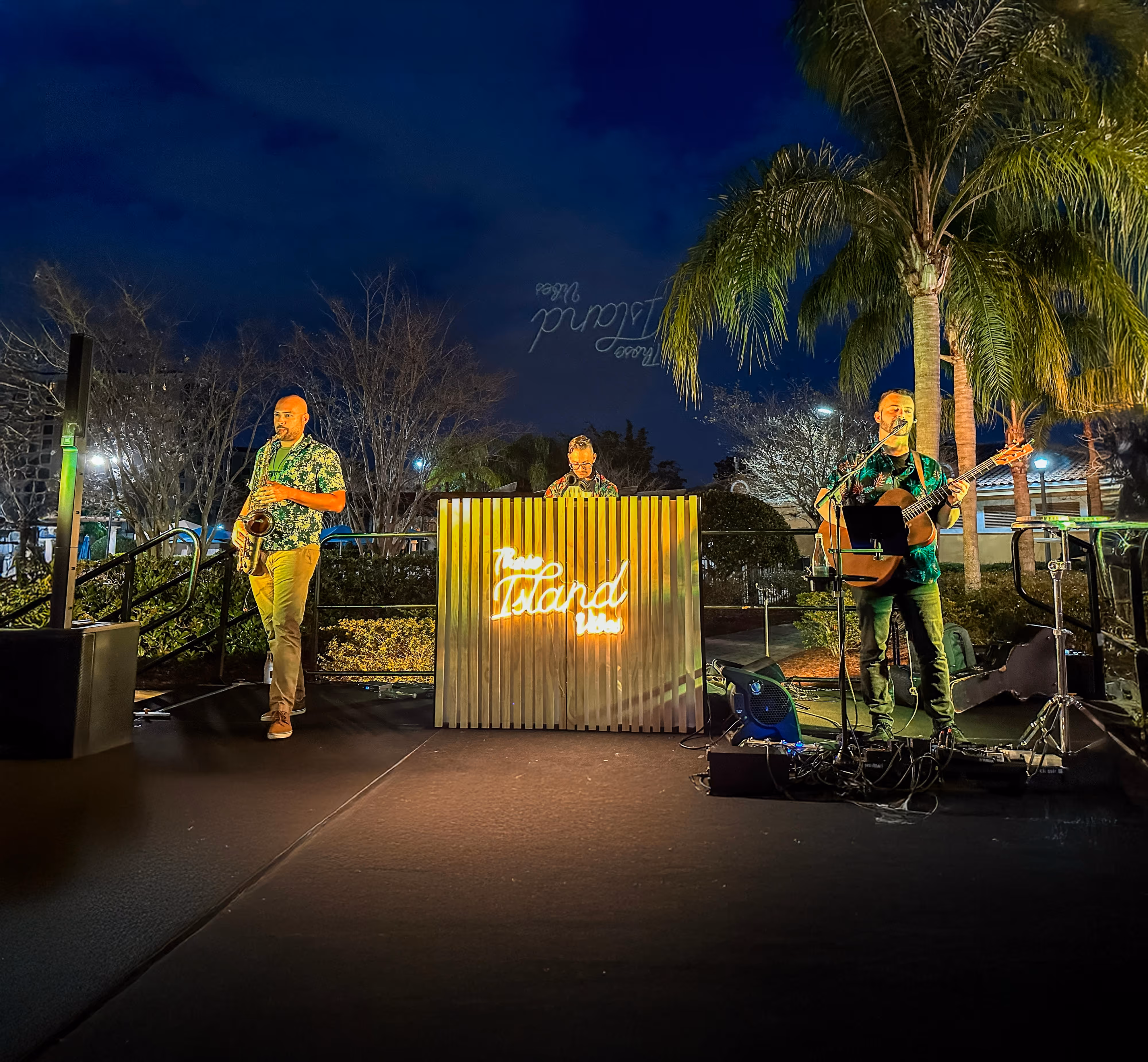 Three musicians performing on an outdoor stage at night with palm trees and bare trees in the background; one plays saxophone, one stands behind a wooden DJ booth with neon 'Island' sign, and one plays guitar and sings into a microphone.