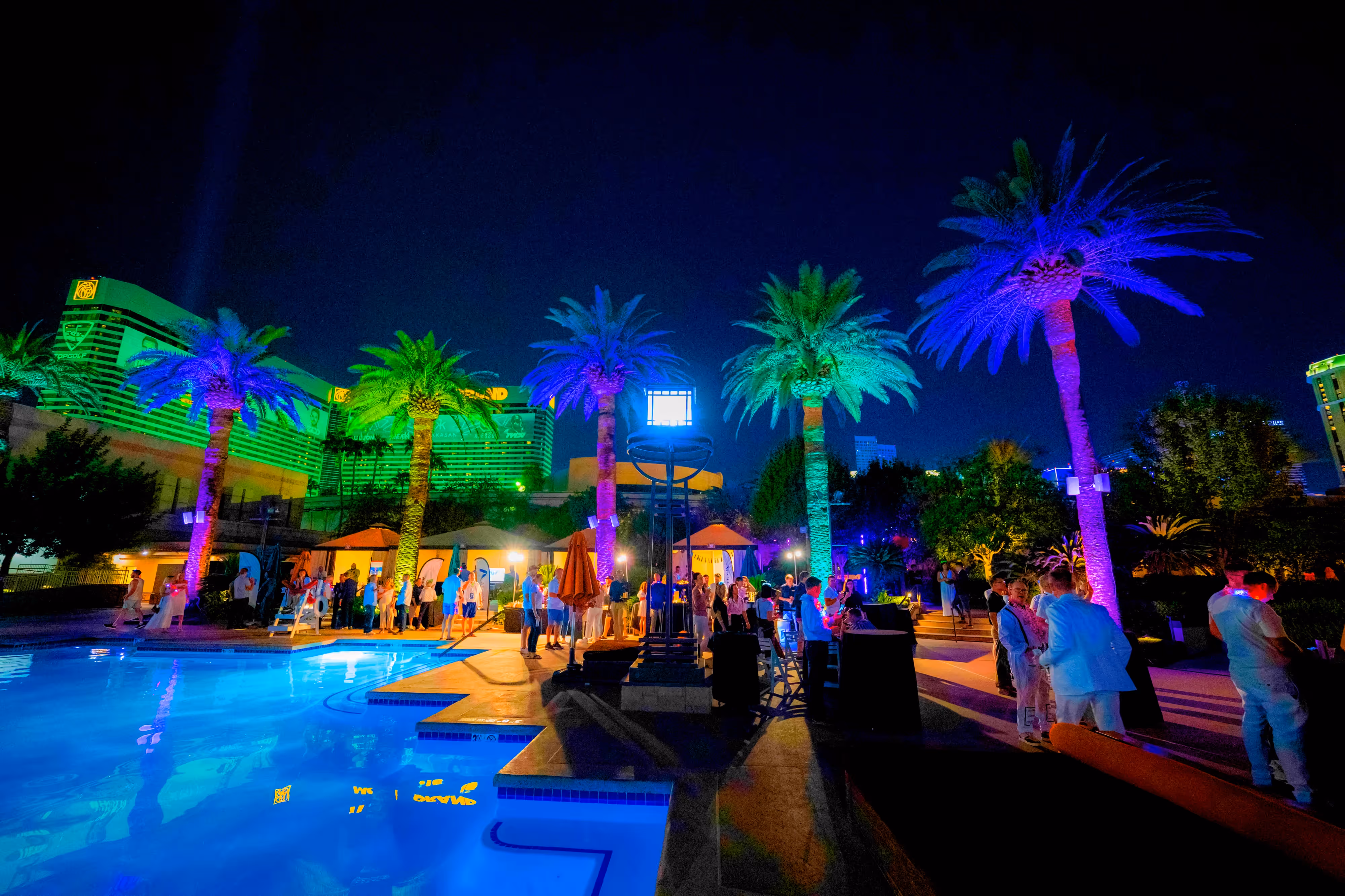 Nighttime poolside scene with people socializing, illuminated palm trees, and bright city lights in the background.