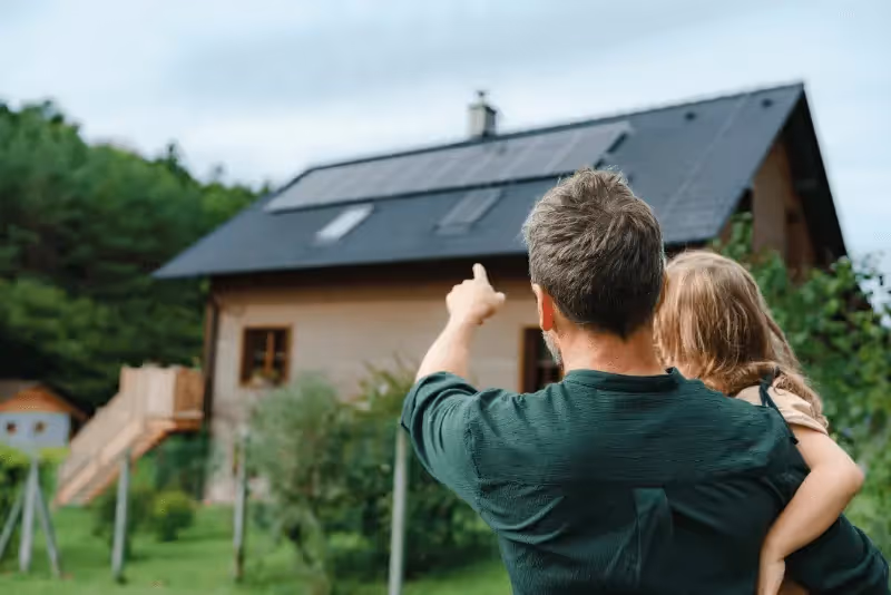 Image of a man holding a small child pointing at solar panels on the roof of a house
