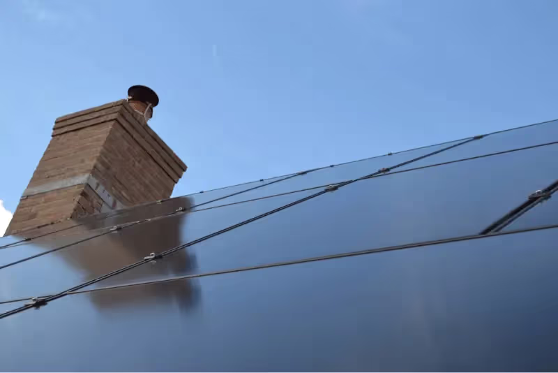 Picture of all black solar panels on a roof of a residential house with a chimney in the background.