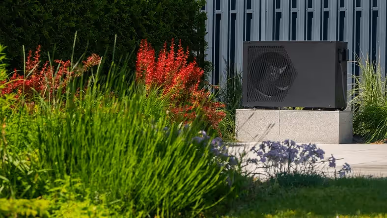 A heat pump on a plinth in a garden near a fence, with plants around