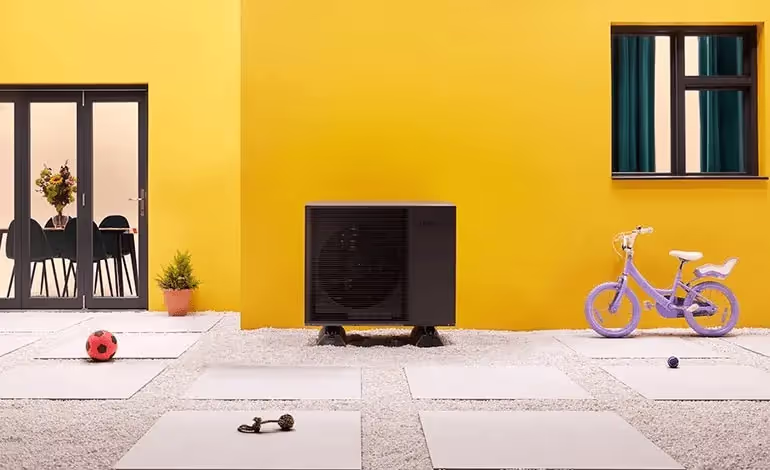 Picture of the Samsung Quiet R32 Heat Pump outside a home with a yellow wall and window into the living room