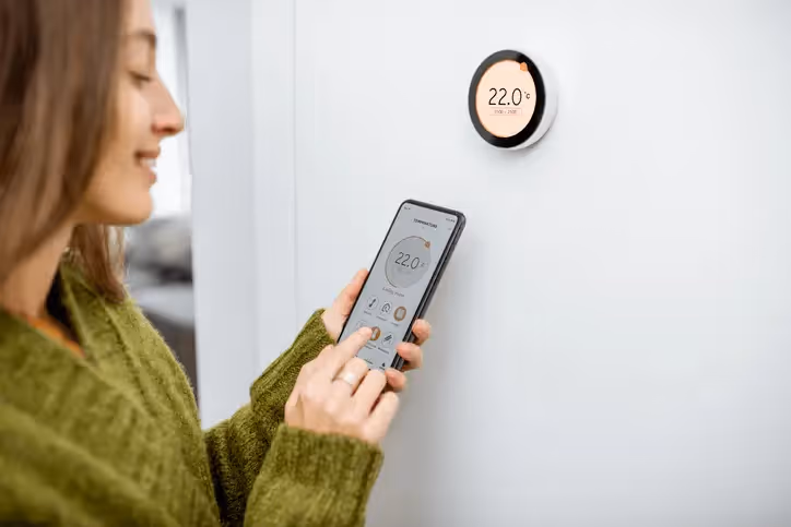 A lady standing infront of a smart thermostat on a wall, with her mobile phone in her hand on a heating app