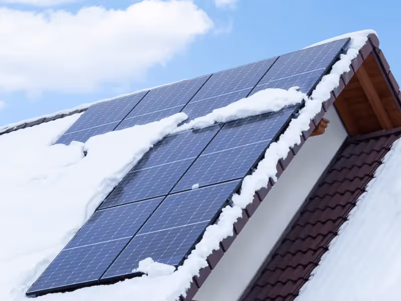 An image of a house with snow on the roof, solar panels sticking our from the snow and a clear blue sky